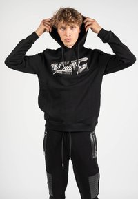 Sudadera negra con un estampado gráfico en el frente y cordones, con puños acanalados. Combinada con pantalones negros que muestran detalles con cremalleras.