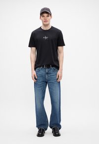 T-shirt de algodão preto com um pequeno logótipo, combinado com jeans de perna reta azul claro e sapatos de couro pretos. Complementa com um boné cinza.