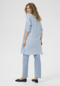 Ljusblå, långärmad skjorta med en texturerad väv, kombinerad med ljusa jeans och svarta platta skor. Modellen ses bakifrån.