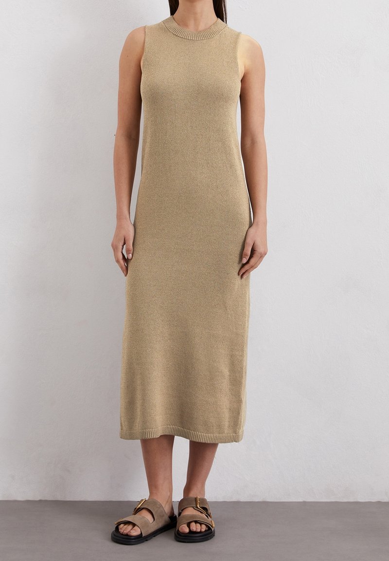 Abito in maglia beige senza maniche con scollo rotondo e rifiniture a coste. L'abito arriva a metà polpaccio ed è abbinato a calzature marroni con dettagli dorati.