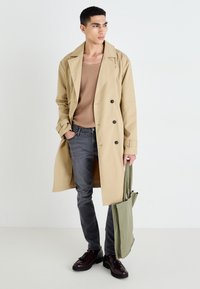 Vaaleanbeiget kaksirivinen trenchcoat, harmaat kapeat farkut, ruskeat kengät ja vihreä kantokassi. Sileä kangas ja jämäkkä muotoilu.