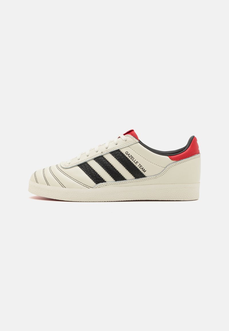 Cream leather sneakers with black stripes, red heel accents, and textured details. Features a rubber sole and lace-up closure.