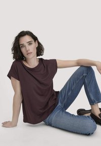 Burgunderfarbendes T-Shirt mit kurzen Ärmeln und lockerem Schnitt, kombiniert mit hellblauen Jeans. Modell sitzt, trägt dunkle Slipper. Neutraler Hintergrund.