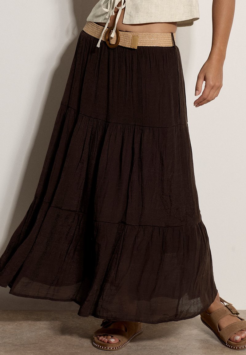 Femme portant une longue jupe noire à volants avec une ceinture beige, un crop top beige et des sandales marron, debout contre un mur uni.