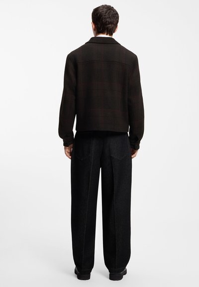 Homme portant une veste à carreaux sombre et un pantalon noir ample, debout de dos sur un fond blanc uni.