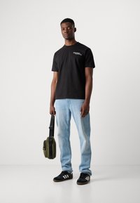 Black short-sleeve t-shirt with white text, light blue jeans, black sneakers with white stripes, and a green crossbody bag.