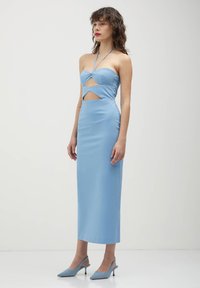 Frau, die ein hellblaues schulterfreies Midikleid mit geometrischen Ausschnitten und passenden spitzen Slingback-Absätzen trägt, vor einem schlichten Hintergrund steht.