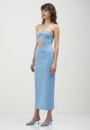 Frau, die ein hellblaues schulterfreies Midikleid mit geometrischen Ausschnitten und passenden spitzen Slingback-Absätzen trägt, vor einem schlichten Hintergrund steht.