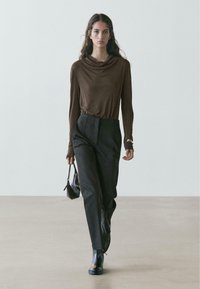 Brązowy top z golfem i długimi rękawami, zestawiony z wysokim stanem czarnych spodni. Modelka niesie małą czarną torebkę, nosi czarne botki za kostkę.