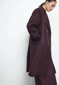 Langer burgunderroter Wollmantel mit doppelt geknöpftem Verschluss, breiten Revers und Seitentaschen. Mit lockerem Schnitt und mittellangem Design.