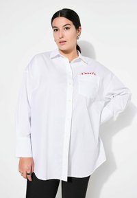 Camicia bianca con bottoni, dotata di taschino sul lato sinistro con testo rosso che dice "Don't." La camicia ha una vestibilità ampia e maniche lunghe.