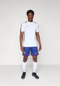 Nike white athletic shirt with black shoulder accents, blue Barcelona shorts featuring a logo, white socks, and black cleats.