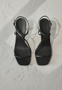 Schwarze Sandalen mit Absatz, dünnen, kreuzweise angeordneten Riemen und einem quadratischen Zehenbereich. Glatte, matte Textur mit flacher Sohle. Keine sichtbaren Beschläge oder Verzierungen.