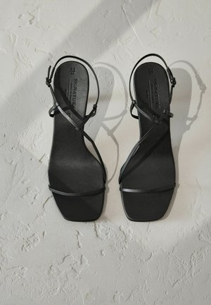 Sorte hæle sandaler med tynde, krydsende stropper og firkantet tå-design. Glat mat overflade med en flad sål. Ingen synlig hardware eller udsmykninger.