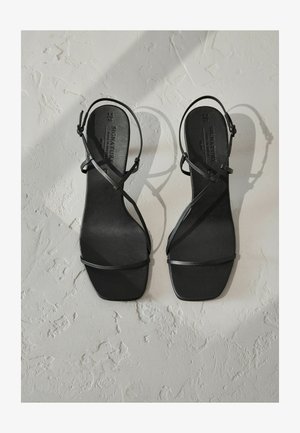 Sorte hæle sandaler med tynde, krydsende stropper og firkantet tå-design. Glat mat overflade med en flad sål. Ingen synlig hardware eller udsmykninger.