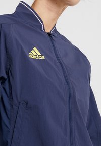Woman wearing a navy blue Adidas zip-up jacket with a yellow logo on the chest and a white-striped collar, facing slightly right.