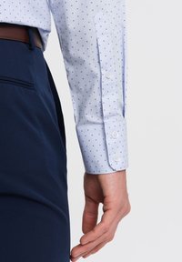 Camicia bianca con un motivo a piccole x blu, con polsini a bottoni, abbinata a pantaloni blu navy. La camicia presenta una tasca appuntita e una trama del tessuto liscia.