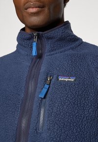 Navy fleece jacket with a high collar, textured fabric, and a zippered front. Features a blue zipper pull and a Patagonia logo on the chest.
