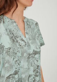 Vaalean mintunvihreä paita, jossa on V-pääntie, lyhyet hihat ja luontoteemainen mustavalkoinen kukkakuosi. Mukana on pystysuora pleat-yksityiskohta.