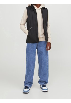 Personne portant une veste noire sans manches zippée sur un sweat à capuche beige, un jean bleu à jambes larges et des baskets blanches avec des touches bleues, debout devant un fond uni.