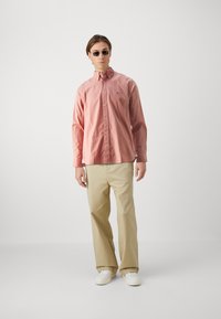Camicia button-up rosa chiaro, tessuto di cotone, maniche lunghe e design aderente. Pantaloni beige, stile casual, abbinati a sneaker bianche.