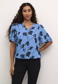 Blaue Blumenbluse mit V-Ausschnitt, kurzen Ärmeln und schwarzen Blumenmustern auf einem hellblauen Hintergrund, kombiniert mit schwarzen Hosen.