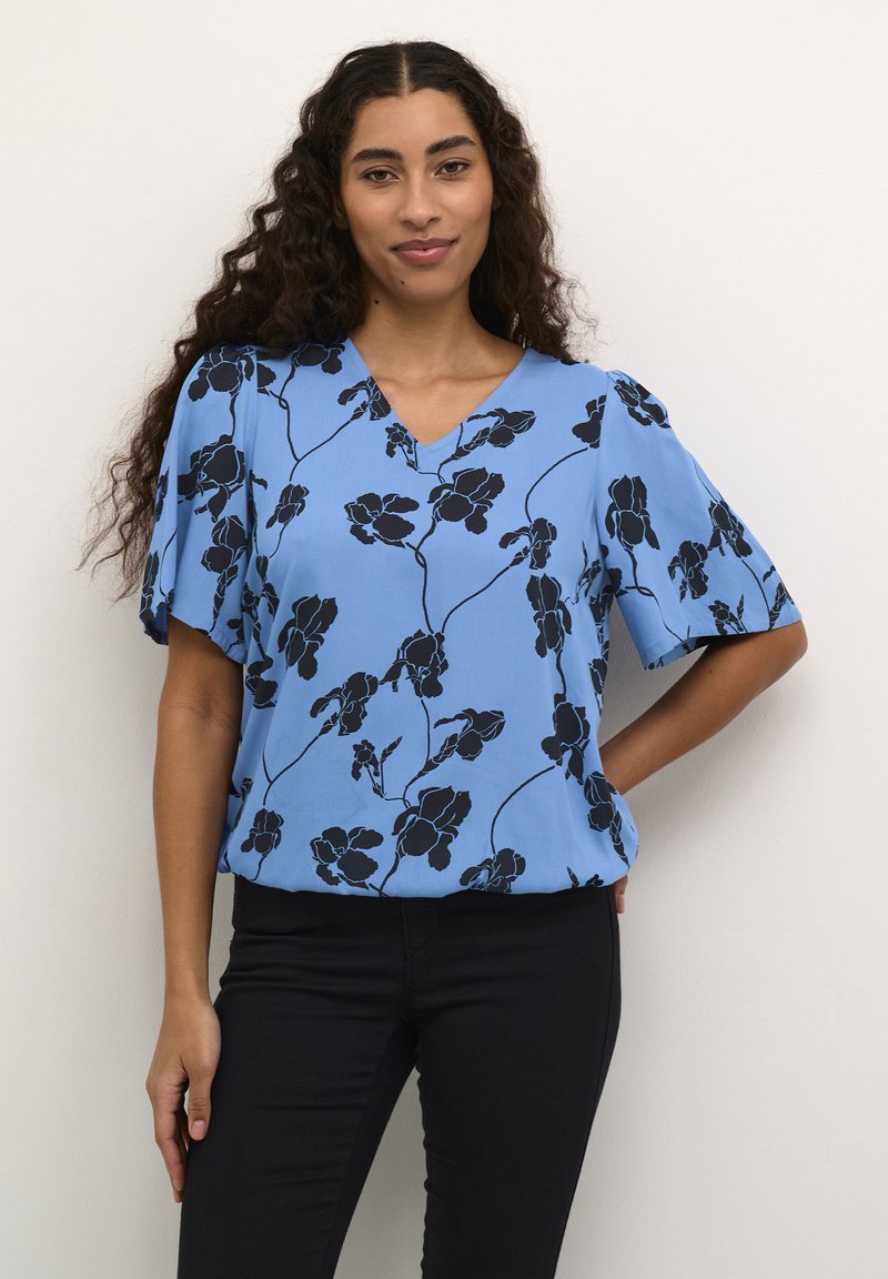 Blaue Blumenbluse mit V-Ausschnitt, kurzen Ärmeln und schwarzen Blumenmustern auf einem hellblauen Hintergrund, kombiniert mit schwarzen Hosen.