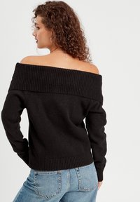 Sort striktrøje med off-the-shoulder design, ribbet halsudskæring og manchetter, parret med blå højtrukne jeans, der viser en afslappet pasform og tekstur.