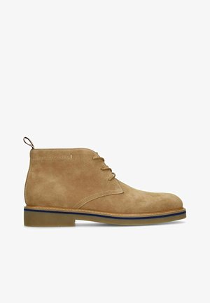 Tan suede ankle boot with three eyelet laces, blue-striped sole, and pull tab at the back.