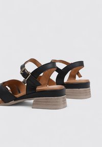 Sandales en cuir noir avec des brides de cheville réglables, un petit talon carré et une semelle légère marron à motifs.