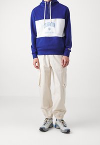 Niebieska i biała bluza z kapturem z logo "Champion", w połączeniu z kremowymi spodniami cargo z bocznymi kieszeniami i elastycznymi mankietami, noszona z szarymi trampkami.