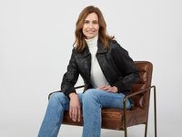 Schwarze Lederjacke mit durchgehendem Reißverschluss, weißer Rollkragenpullover und blaue Schlagjeans. Sitzt auf einem bronzeumrahmten Stuhl mit braunem Lederbezug.