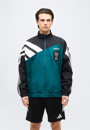 Ung mand iført teal og sort Adidas Liverpool fodboldjakke og sorte Adidas shorts, stående mod en ensfarvet hvid baggrund.