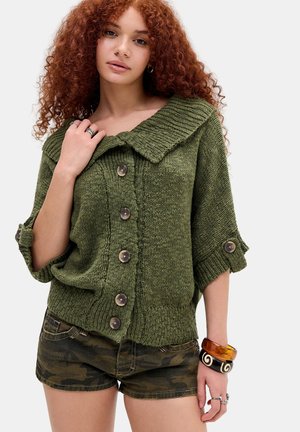 Junge Frau mit lockigem roten Haar, die einen grünen, geknöpften Strickpullover mit hochgekrempelten Ärmeln und Tarnshorts trägt, ergänzt durch Armbänder und Ringe.
