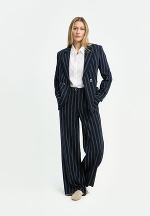 Frau trägt einen dunkelblauen Nadelstreifenanzug mit weitem Bein und ein weißes Hemd mit Knöpfen, steht mit den Händen in den Taschen vor weißem Hintergrund.