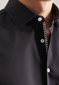 Camisa oscura con un cuello estructurado, que presenta botones blancos y una tapeta con patrón. Tejido suave con un ligero brillo.