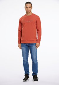 Felpa color ruggine con scollo rotondo, con la scritta "COPENHAGEN"; abbinata a jeans blu e scarpe sportive nere.