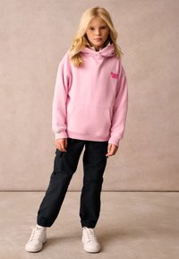 Felpa in pile rosa con tasca frontale e logo ricamato, abbinata a pantaloni cargo neri. Sneakers bianche completano l'outfit.