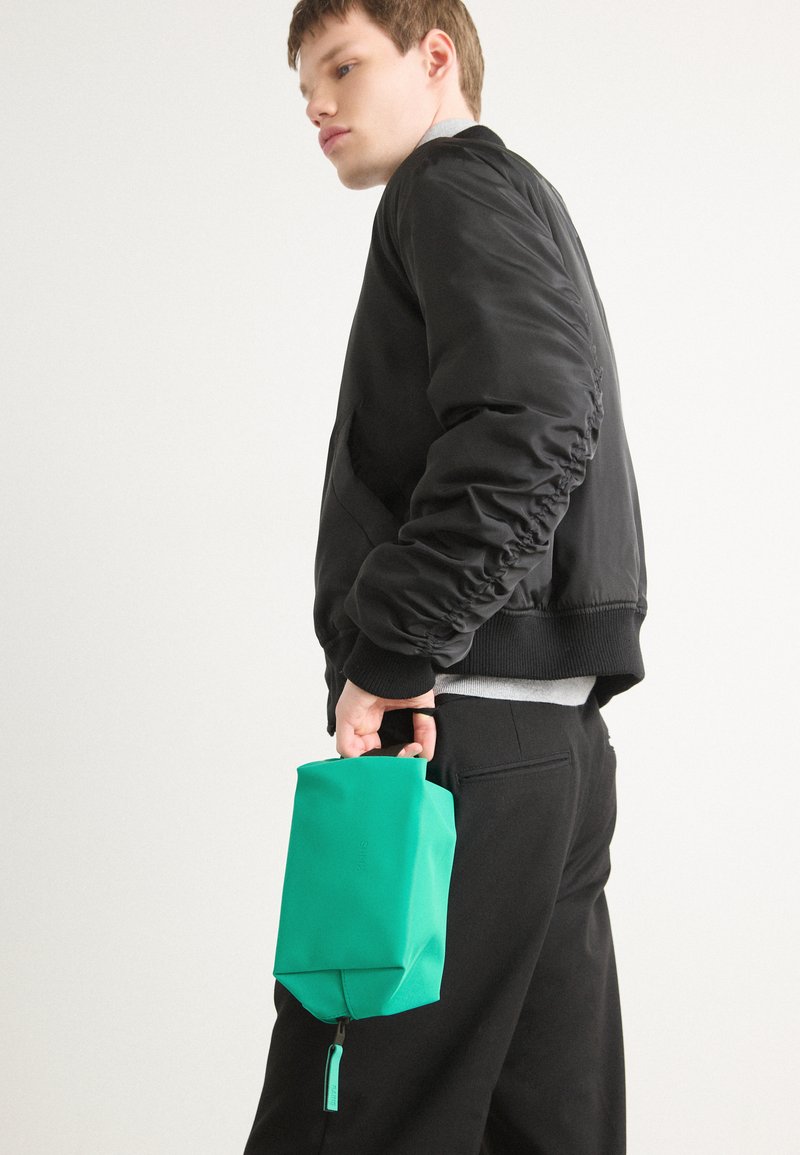 Borsa verde con zip realizzata in materiale testurizzato, tenuta in una mano. Abbinata a una giacca bomber nera e pantaloni neri. Design semplice e moderno.