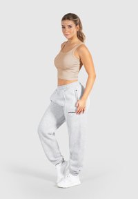 Donna in canotta beige a croppata, pantaloni grigi con la scritta "Sailbuddy" e sneakers bianche su uno sfondo chiaro uniforme.