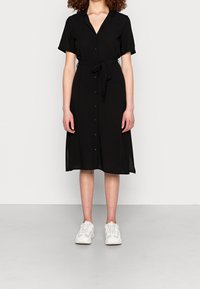 Abito nero con bottoni, maniche corte e vita regolabile, realizzato in tessuto leggero, lungo fino al ginocchio, abbinato a sneaker bianche.