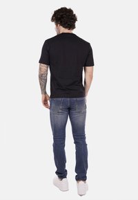 T-shirt nero a maniche corte, jeans in denim blu e sneaker bianche. I jeans hanno una vestibilità classica con tasche posteriori. Vista posteriore.