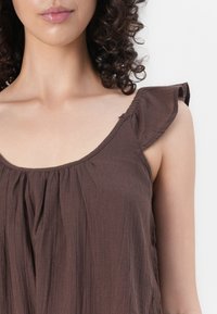 Femme aux cheveux bouclés portant un haut marron sans manches avec un détail froncé à l'épaule sur un fond uni.