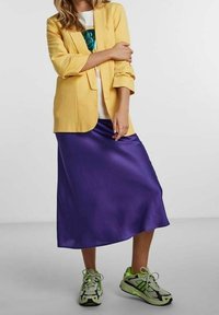 Żółta marynarka, biała koszulka z grafiką, purpurowa satynowa spódnica oraz zielono-czarne sportowe buty, tworząc warstwowy, casualowy look.