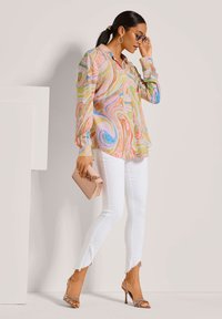 Buntes Langarmshirt mit wirbelndem Muster in Pastellfarben, kombiniert mit weißen, zerrissenen Jeans und nudefarbenen High Heels. Hält eine kleine pinke Clutch.