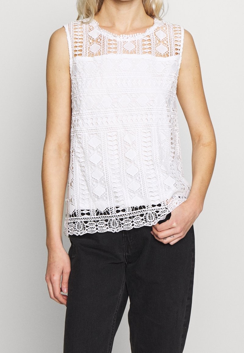 Vit ärmlös topp med intrikata spetsmönster och scalloped nederkant. Texturerad design med rund halsringning och öppna stickade inslag.