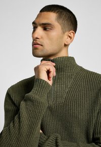 Tikkitakkipaita tummassa oliivinvihreässä, jossa on korkea kaulus, ribboitunut pinta, osittainen vetoketju ja leveät hihansuut. Yksityiskohtainen ompelu edessä.