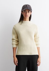 Cremefarbener Waffelstrickpullover mit hohem Stehkragen und gerippten Bündchen, kombiniert mit einer schwarz-grau karierten Mütze. Getragene schwarze Hose.