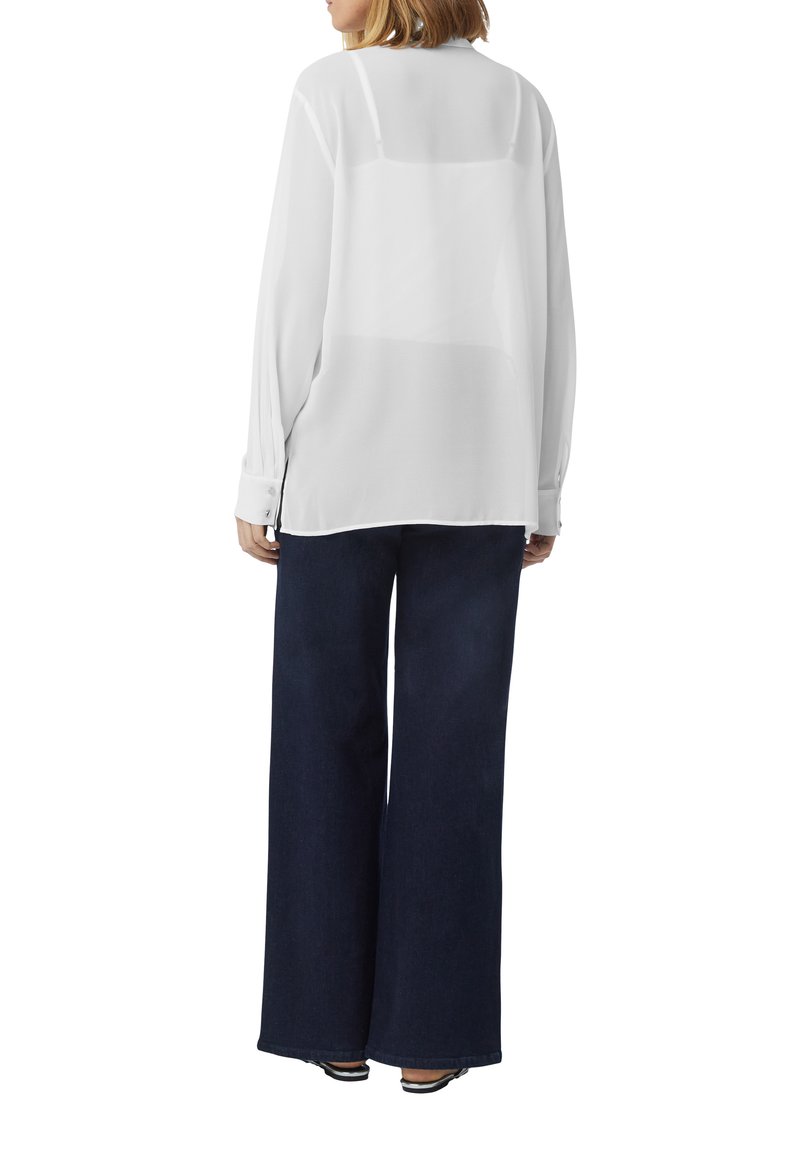 Weiße, transparente Bluse mit langen Ärmeln und lockerem Schnitt. Kombiniert mit dunkelblauen, weit geschnittenen Jeans. Verfügt über geknöpfte Manschetten und Seitenschlitze.