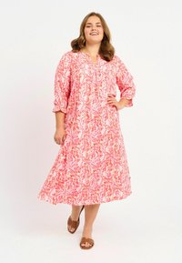 Fließendes rosa Kleid mit wellenförmigen abstrakten Mustern, rundem Ausschnitt und Dreiviertelärmel. Das Model trägt braune Sandalen und zeigt einen lässigen Schnitt.